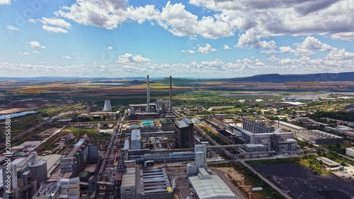 The scene shows a wide view of an industrial area featuring several large buildings and smokestacks. Green fields stretch out in the background under a clear blue sky with fluffy clouds.