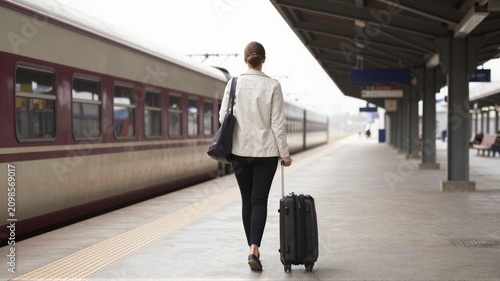 Woman walking along railway platform with rolling suitcase and shoulder bag beside stopped train carriages, preparing for journey under station canopy