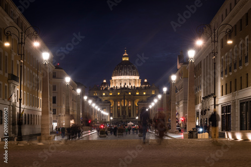 Symmetrical night view of the illuminated St. Peter's Basilica along Via della Conciliazione with motion blurred people in Rome, Italy