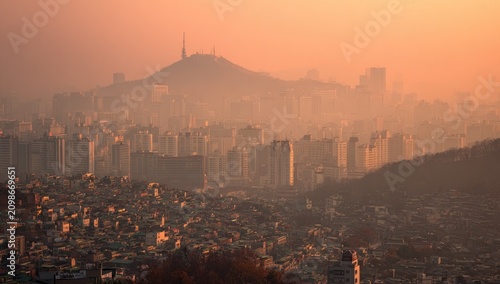 Elevated cityscape under orange hazy sky, distant mountain & tower. Buildings blanket the urban sprawl