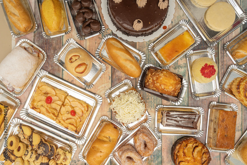 Canvas Print Top-down view of assorted baked goods like chocolate cake, apple pastries, bread rolls, and cookies arranged on silver trays over a wooden surface