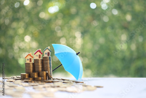 Miniature houses on stacked gold coins protected by a blue umbrella, concept of real estate insurance, property protection and financial security for home investment.