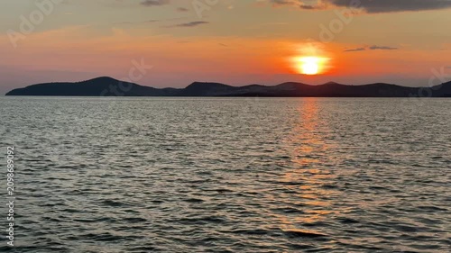 A calm, warm sunset over the Aegean Sea in Gulluk, Turkey. The sun hangs low in a hazy orange sky, casting a bright golden light path across the gently rippling water toward distant dark hills.