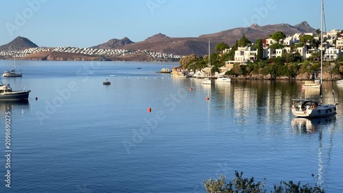 Scenic view of the calm, blue bay in Bitez, Turkey, with luxury boats anchored on the still water near a hillside resort town under a clear morning sky.