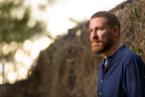 Portrait of a middle-aged man with a beard looking away, wearing a navy blue shirt standing against a rustic natural background with copy space.