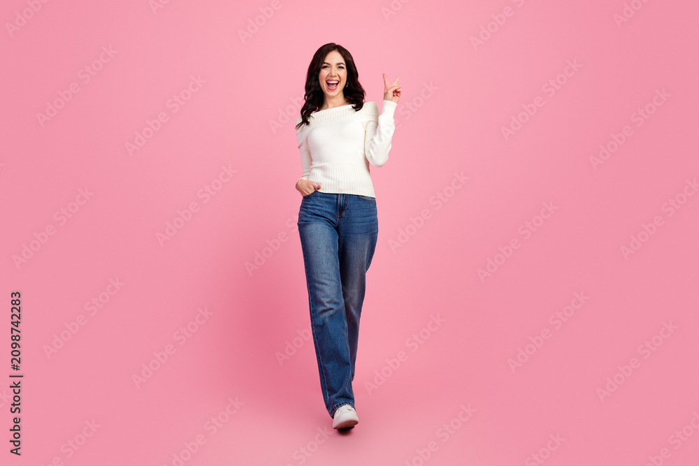 Obraz premium Happy young woman walking and flashing a peace sign in casual jeans and white sweater on a pink background, confident lifestyle portrait and fashion studio shot