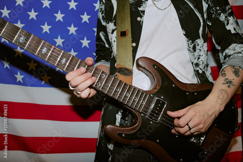 Close up of tattoed musician playing electric guitar at home with American flag backdrop, expressing rock identity and performance culture. Ideal for music marketing and artist branding