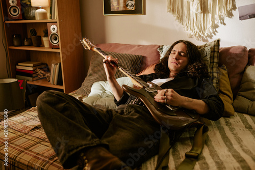 Fotografía Young adult musician lying on bed, playing electric guitar at home, practicing songwriting and creative performance