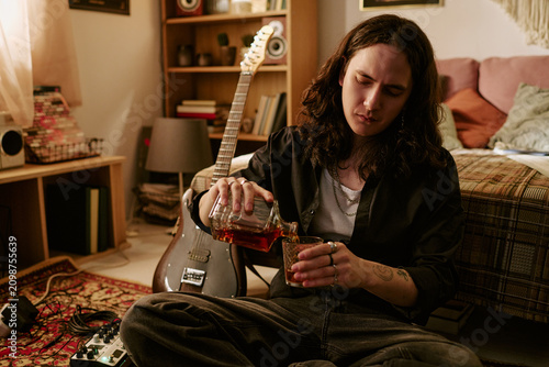 Fotografie Young adult musician sitting on floor at home, pouring whiskey into glass near electric guitar, expressing creative burnout and substance use
