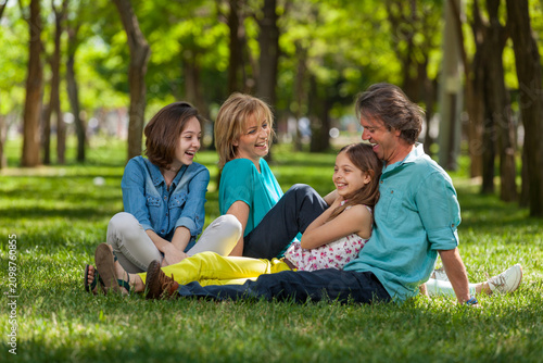Happy family sitting on the grass in a sunny park, candid portrait of parents with daughters, outdoor leisure, and family bonding concept.