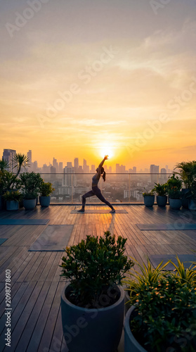 Woman performs yoga pose on rooftop at sunset with city skyline view. Warm colors fill the sky, creating a tranquil scene. Concept of fitness, wellness, lifestyle