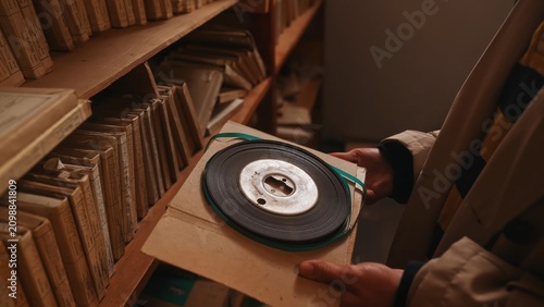 Hands hold a vintage audio reel on a cardboard sleeve between archive shelves. Concept of media history, research, media preservation, catalog library, documentary content and nostalgia.