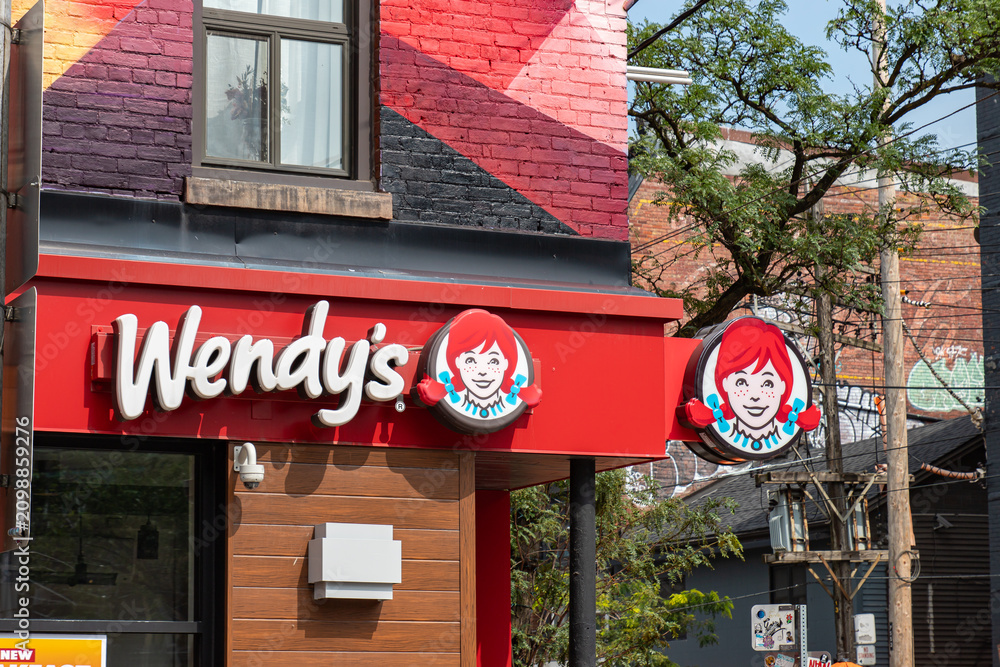 Fototapeta premium Toronto, ON, CANADA - Aug 8 2024 : Exterior of a Wendy's restaurant with the red logo signage and Wendy mascot circle signs mounted on a red awning below a brick building facade.