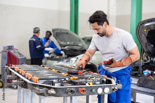 Auto technician servicing an electric car battery pack on a workbench in a garage, with vehicles and coworkers in the background during diagnostics and repair.