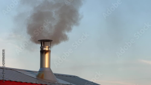 Smoke rises from a metal chimney on a red wooden roof against a cloudy sky. heating, winter home, cozy rural life.