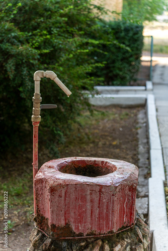Konstfotografi An unusual, rustic outdoor drinking water fountain styled as a giant red hex nut with a metal pipe standing against a backdrop of green bushes and a concrete walkway