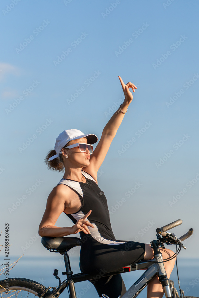 Fototapeta premium Cyclist woman point, outdoor sport training posing with bicycle at sunset showing a relaxed lifestyle during sunny summer outdoor vacation