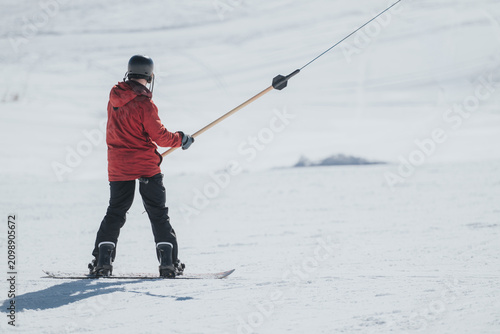 Bilde på lerret A snowboarder wearing a red jacket and helmet holds a tow bar while being pulled across a snow slope