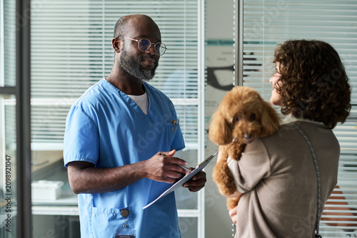 Fotografie Medium shot of Black male veterinarian consulting female pet owner, holding clipboard while examining poodle in clinic