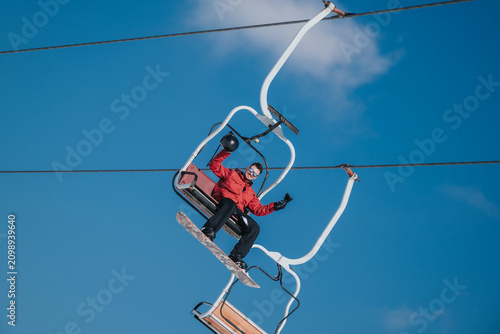Canvas Print A snowboarder sits on a chairlift wearing a red jacket and holding a helmet