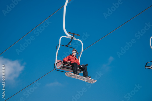 A snowboarder in a red jacket sits on a chairlift holding a snowboard against a clear blue sky. The winter scene shows a rider headed up the mountain on a ski lift.