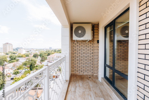 Sunlit balcony with wall mounted air conditioner against beige brick exterior beside sliding glass door, white metal railing, and distant city skyline view from high floor apartment