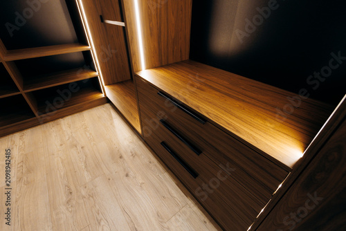 Modern walk-in closet with illuminated wood cabinetry, built-in drawers, open shelving, warm ambient lighting, and light oak flooring in elegant residential interior