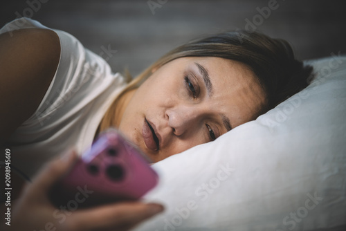 Sleepy woman lying in bed, looking at a clock or phone with a tired expression. Heavy morning awakening, reluctance to get up, and late-night scrolling.