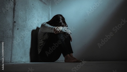 A woman sits alone in a dimly lit corner, hunched over with her head in her hands, conveying a sense of deep sadness and isolation