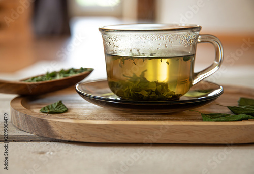 Dried birch leaf powder tea in a glass cup, with Betula leaf powder on a wooden spoon. The glass sits on a home kitchen table, backlit by natural light coming through a window.