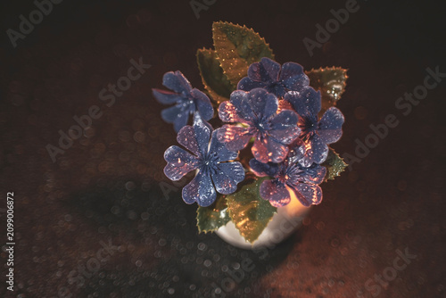 Artificial blue plastic flowers in decorative small pot close up macro photography with warm backlight on dark background with intense round soft focus bokeh effects