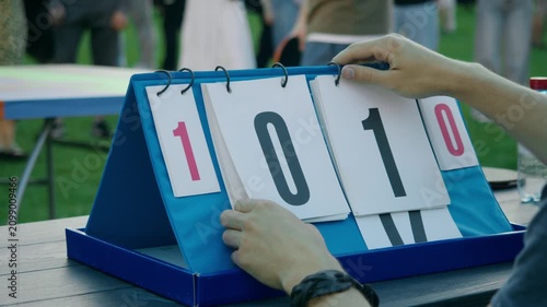 Scorekeeper flips scoreboard card after first point during amateur outdoor table tennis tournament at summer party. Manual scoreboard shows player taking early lead as friends watch exciting ping pong