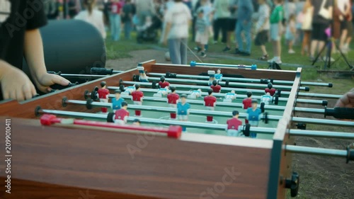 People play table football during lively outdoor summer party with crowd gathering around game. Friends enjoy friendly foosball match as festival atmosphere fills park.