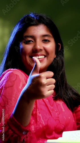 Indian Asian studious teenage beautiful girl sitting at a study table in a cozy lavish home, showing a confident success gesture while studying, writing, demonstrating focus, determination, excellence