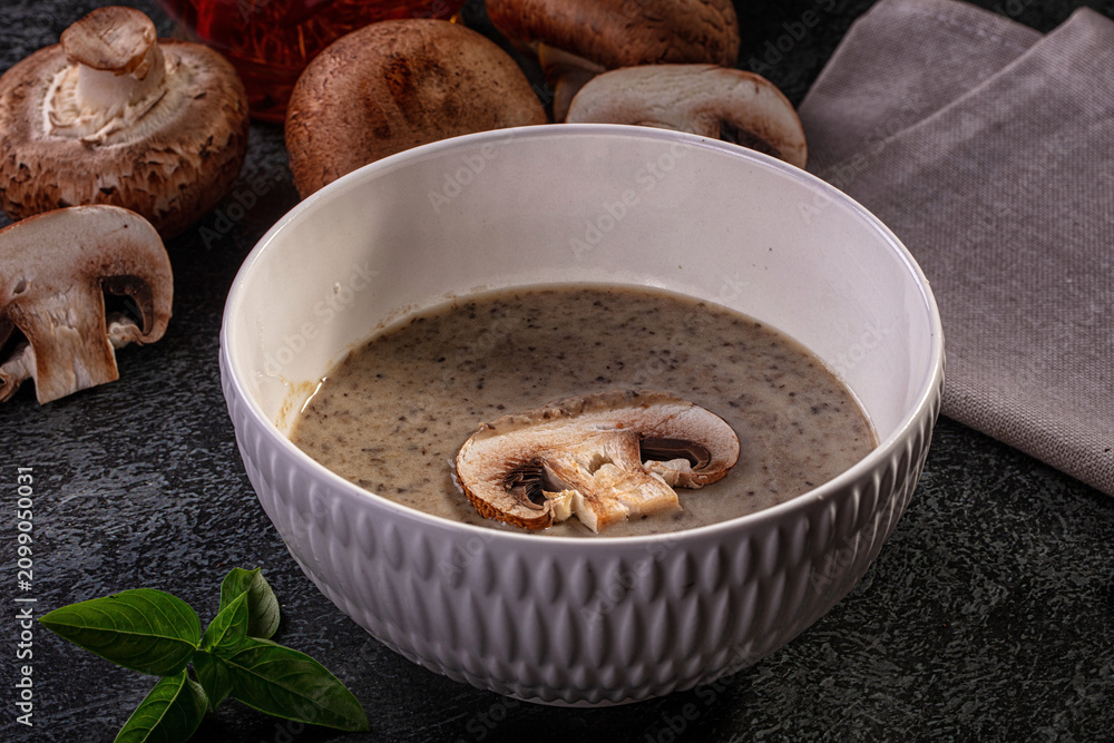 Fototapeta premium Creamy mushroom soup in textured white bowl with fresh mushrooms and basil on dark surface