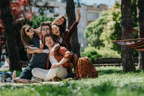 A group of high school students laugh and hug while sitting on a picnic blanket in a park. The young friends pose happily together near trees and a colorful hammock for a relaxed outdoor moment.