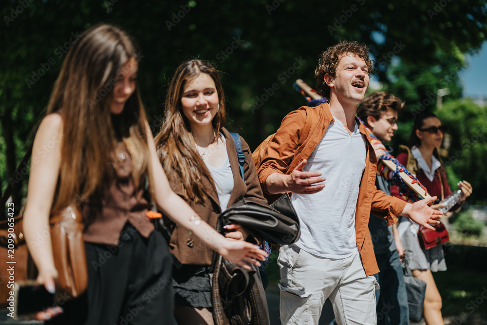 Fototapeta premium High school students walking together outdoors, laughing and chatting. A group of teenagers strolls along a tree-lined path carrying backpacks and bags.