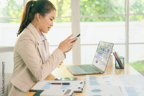 A smiling Asian businesswoman works with financial data charts and a smartphone in the office, analyzing business data graphs.