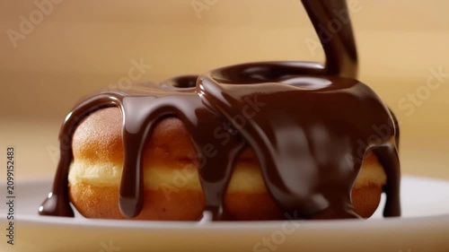 Chocolate syrup drizzles over a freshly baked donut on a white plate, showcasing the smooth flow of chocolate and the soft texture of the pastry