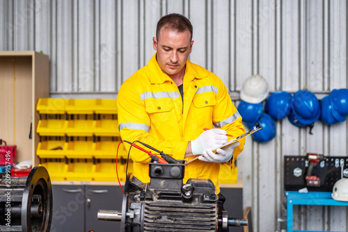 Male industrial electrician in yellow safety jacket inspects an electric motor using a multimeter while writing notes on a clipboard in a factory workshop. Maintenance and quality control concept.