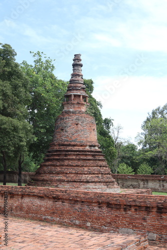 Canvas Print Ancient Buddhist Stupa in Thailand: A weathered brick stupa, a significant religious monument, stands amidst lush greenery and ancient ruins, hinting at a rich cultural past and spiritual serenity