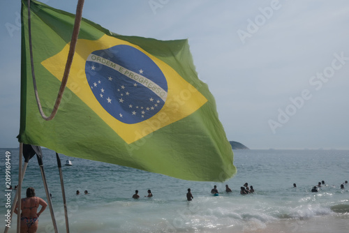 фотографія Bandeira do Brasil tremulando na areia de uma praia no Rio de Janeiro, com o mar azul ao fundo e banhistas na água em um ensolarado dia de verão