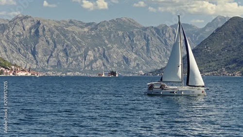 aerial view of a white sailboat under full sail crossing the deep blue water of the bay of kotor, passing the island church of our lady of the rocks and perast below steep mountains, montenegro