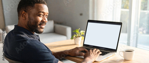 Retrato de perfil de un profesional afroamericano mirando a la cámara junto a su laptop con pantalla blanca para mockup. Concepto de trabajo remoto, tecnología y comunicación digital.