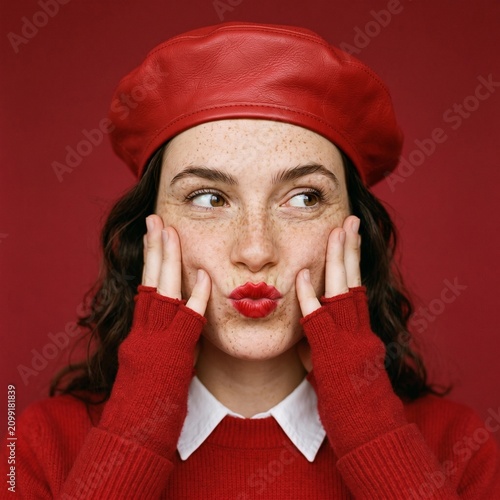 Playful portrait of a freckled young woman puckering her red lips while wearing a vibrant red leather beret and sweater