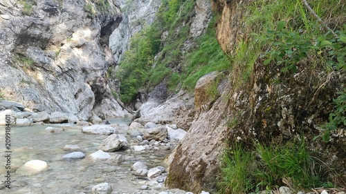 Clear mountain river flows through a dramatic rocky gorge with steep cliffs, mossy stone formations, lush green vegetation, and small boulders on a bright, sunny day in the mountains.