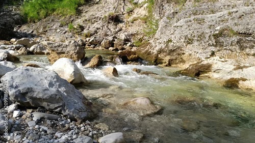 Clear mountain stream with audible rushing water flows over a rocky, boulder-filled riverbed, passing under a steep, grassy and rocky embankment under natural daylight.