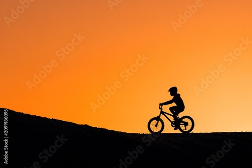 A child rides a bicycle on a hill during sunset. The sky shows bright colors as the sun sets in the background. The silhouette of the child stands out clearly