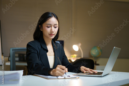 asian businesswoman working at her desk  focused on taking important notes on a notebook while using her laptop  ideal for business and corporate concepts.