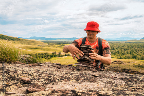 фотографія Man with backpack places tower of flat carne stones on rock outcrop with panoramic view of mountain valley
